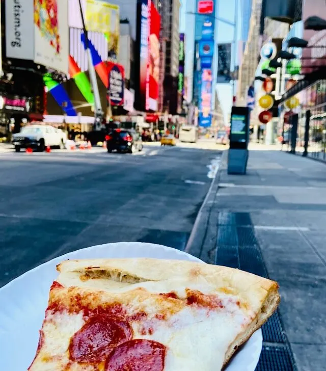 a slice of new york style pepperoni pizza on a white paper plate with the skyscrapers and neon billboards of times square manhattan new york usa in the background