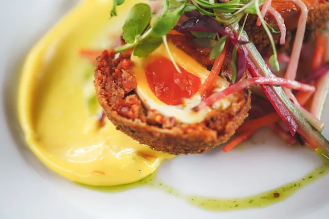half a scotch egg with mustard and arugula on a white plate drizzled with pesto