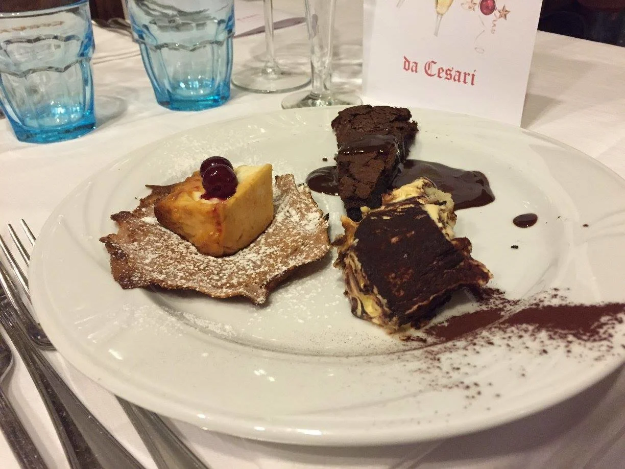 a trio of desserts on a white plate with blue glass tumblers in the background at da cesari restaurant in bologna italy