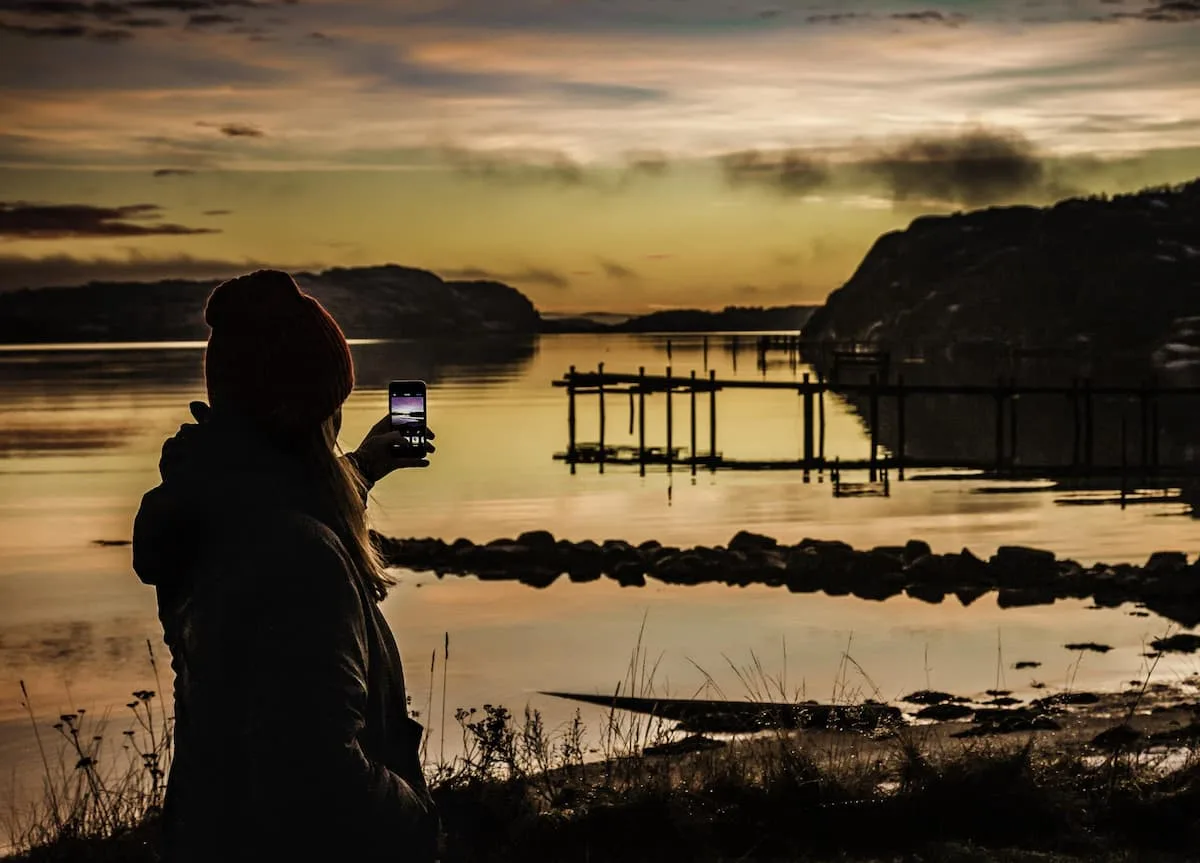 a woman taking a photo of a lake on a smartphone with a wooden pier in the background on a summer night in stockholm sweden