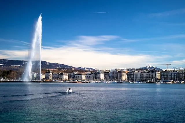 a fountain rising above lake geneva with a boat on it and buildings in the background in geneva switzerland