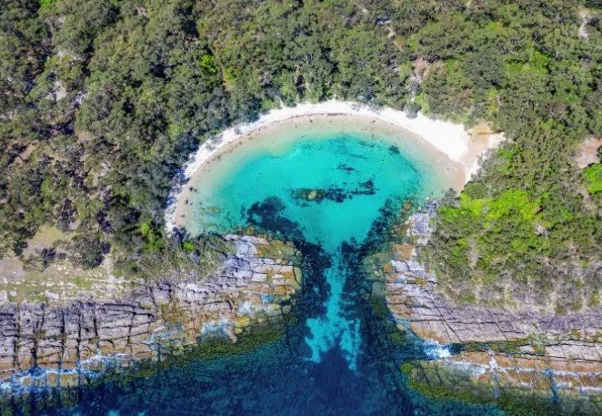 An aquamarine beach with white sand, surrounded by trees, seen straight from above
