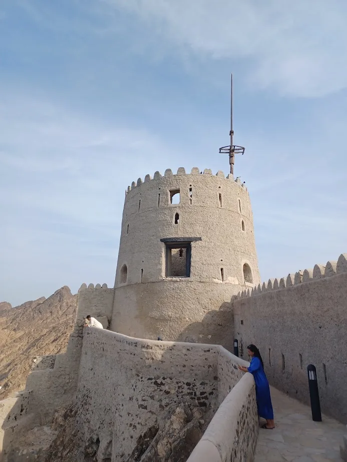 a lady in a blue dress leaning against a wall underneath a tower at mutrah fort muscat oman