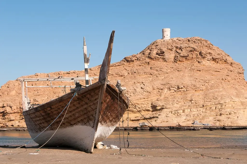 a wooden dhow boat in front of a hillside fort in sur oman