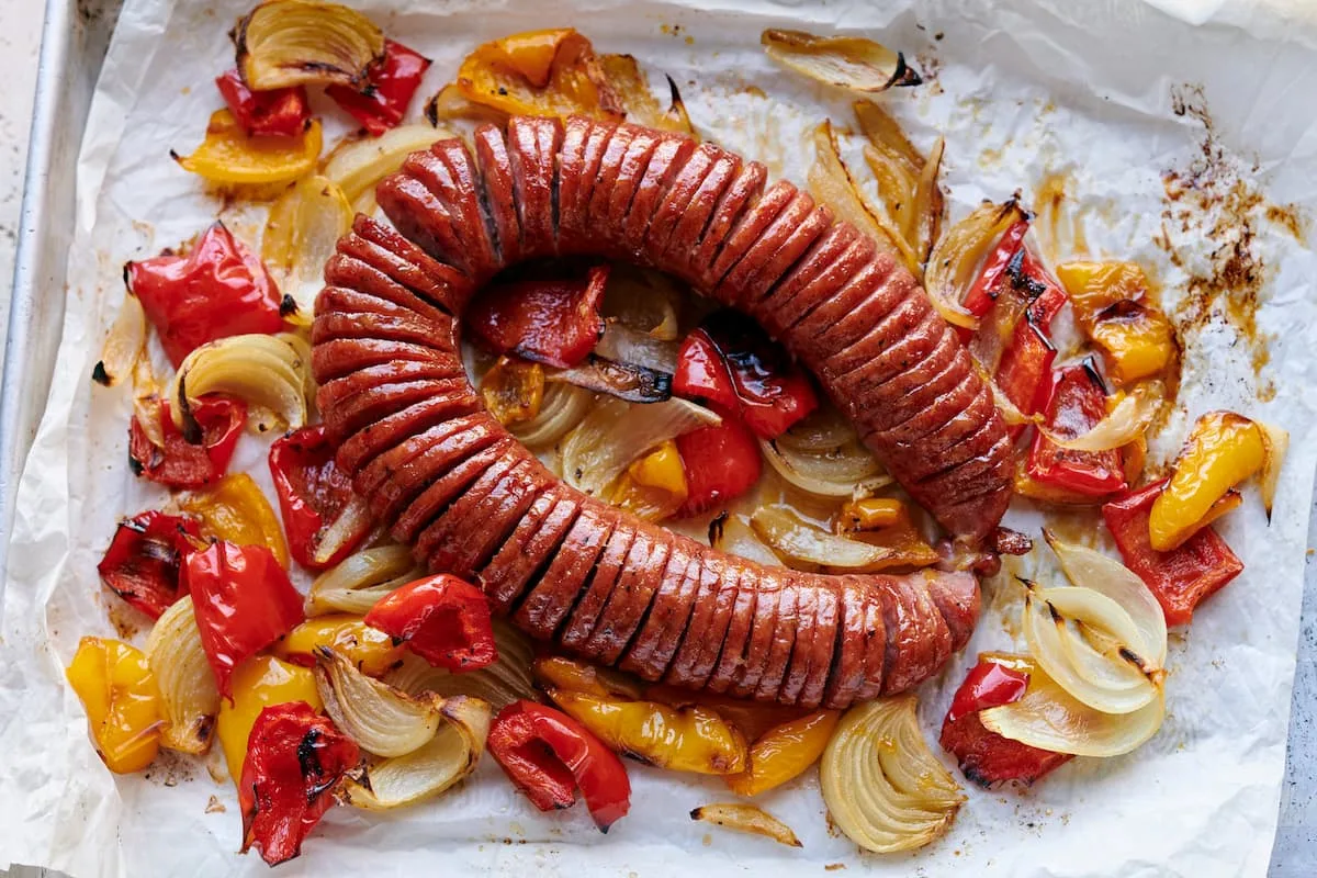 a hassleback kielbasa polish sausage on a baking tray surrounded by red and yellow peppers