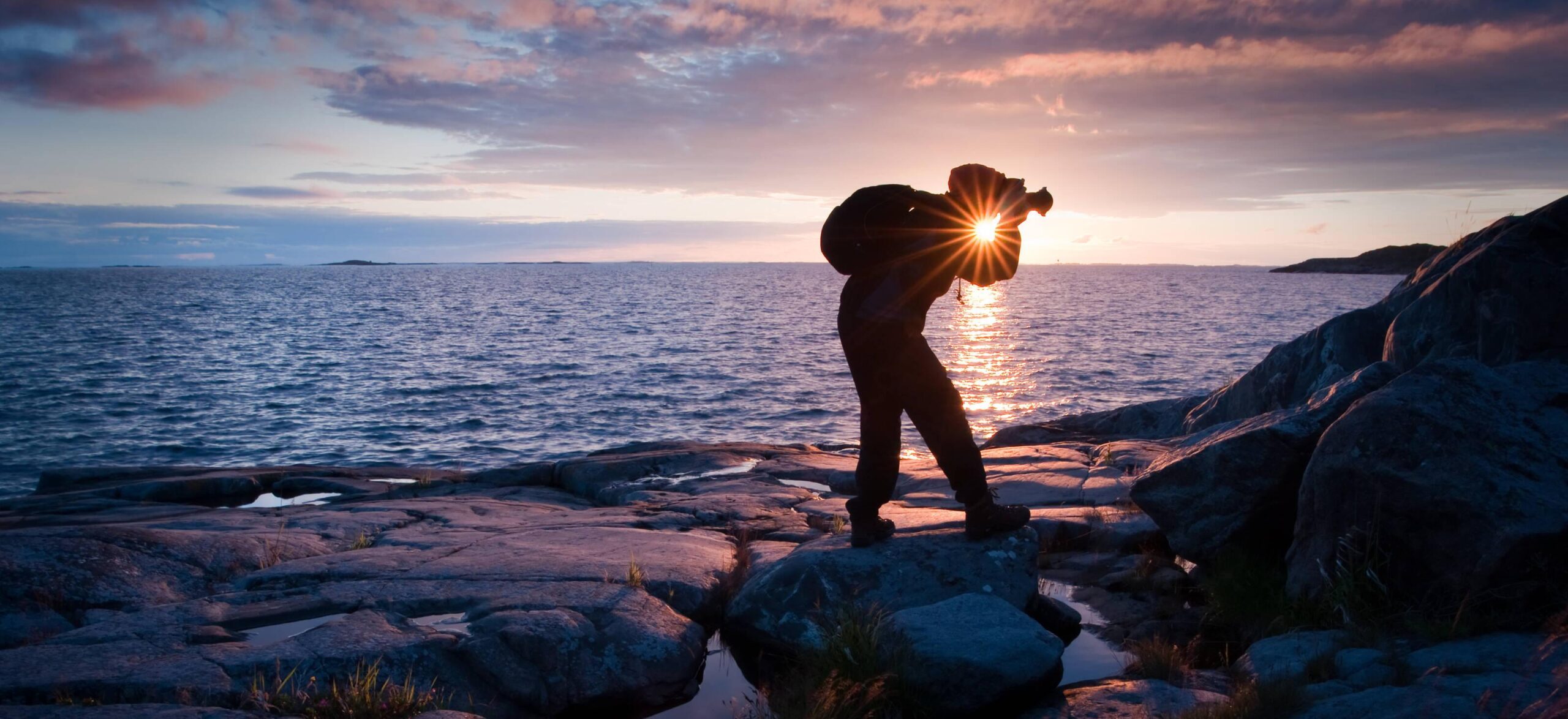 A person with camera walking on rocks by the sea at sunset