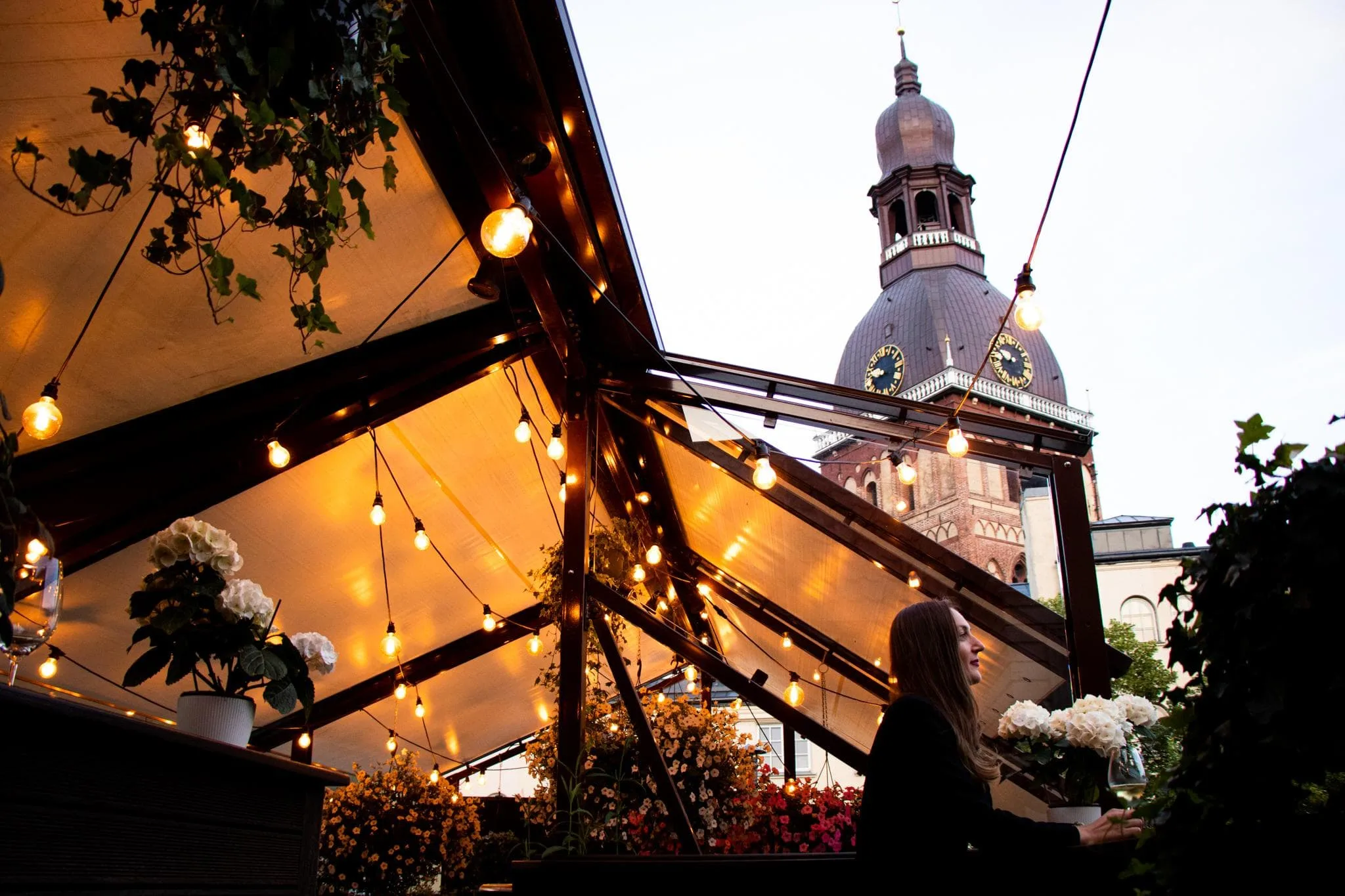 a church tower behind a restaurant awning with fairy lights at le dome restaurant in riga latvia