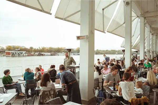 people sit outside a restaurant with tables and a white awning overlooking the Sava river in Belgrade serbia