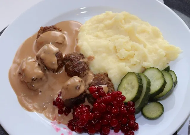 a white plate with swedish meatballs in whisky cream sauce with pickled cucumbers, lingonberries and mashed potato