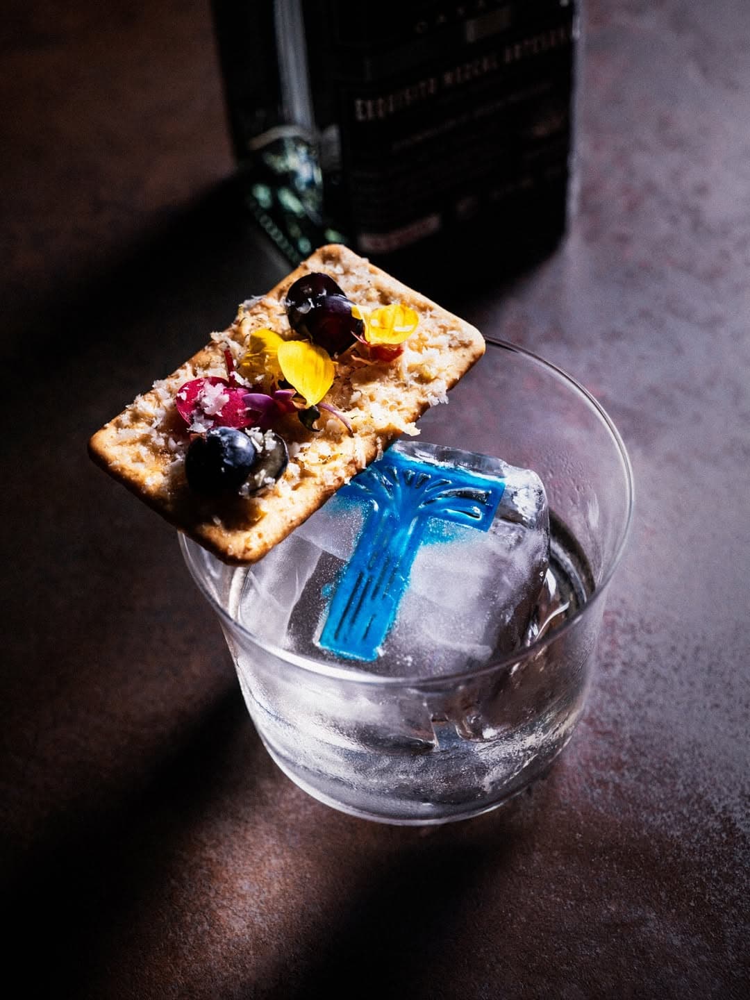Cocktail topped with cracker and edible flowers and served with illustrated ice blocks