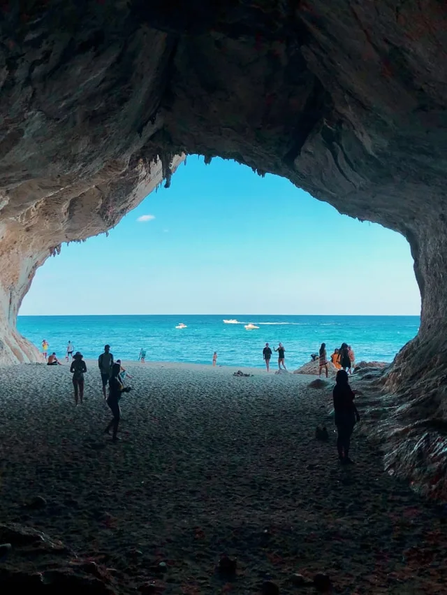 people standing on a pebble beach at the mouth of a cave in sardinia italy