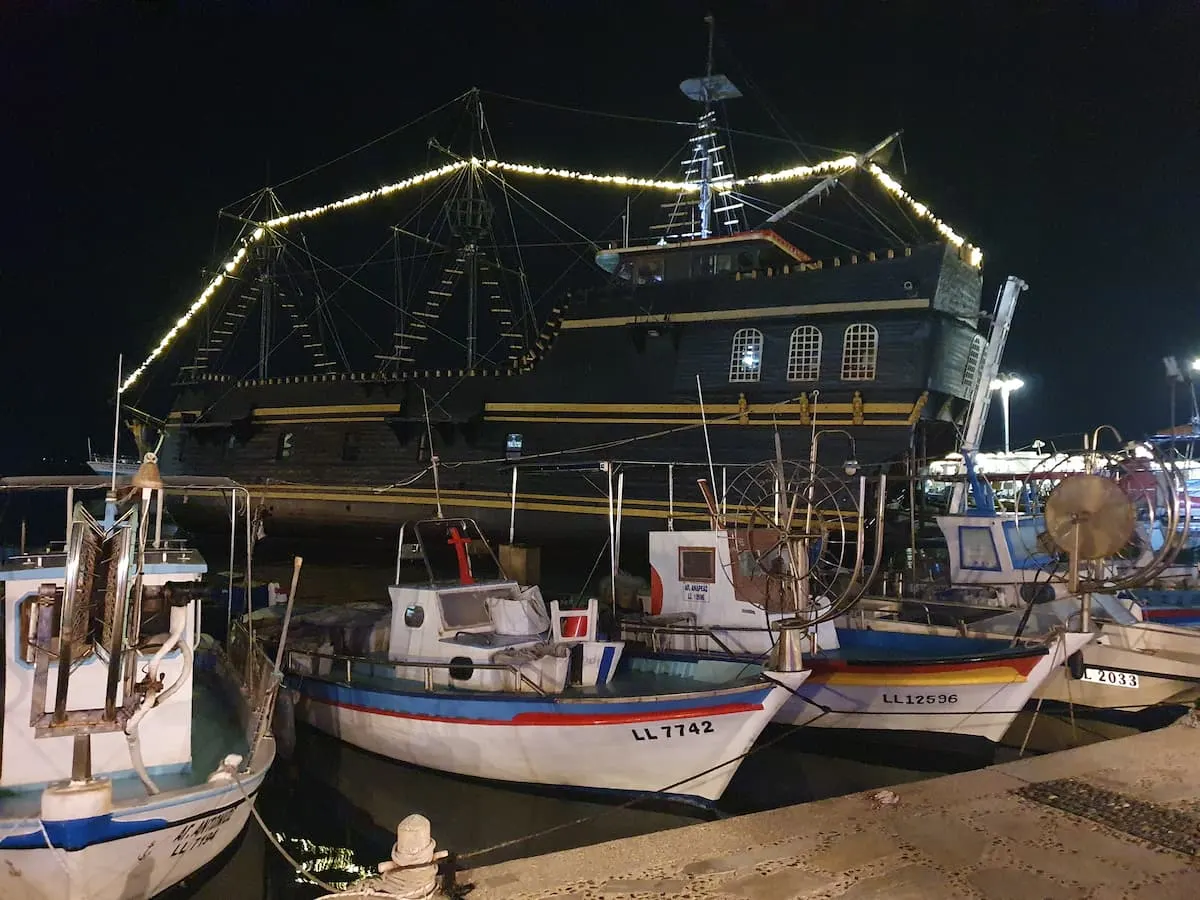 small fishing boats with a large black mock pirate ship in the background in aiya napa harbor on the mediterranean ocean in cyprus 