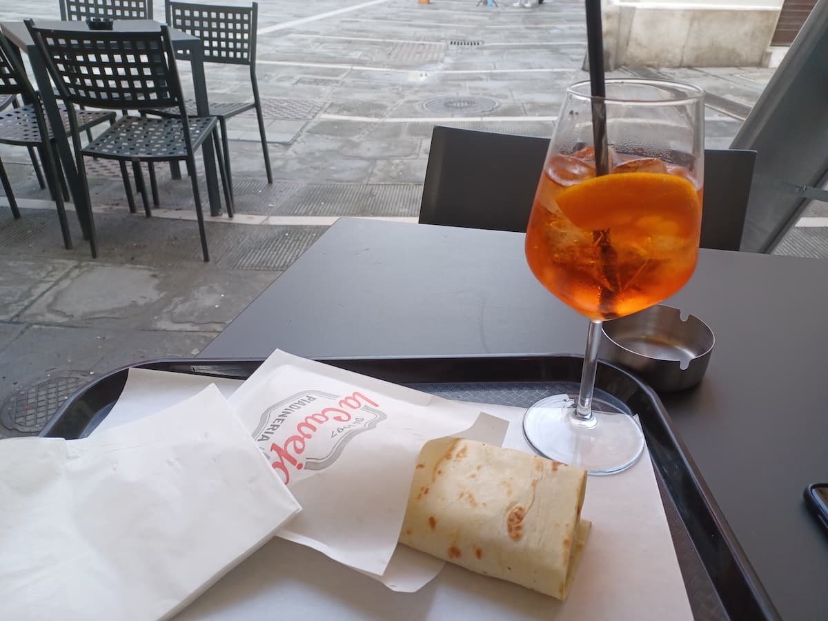 an aperol spritz in a glass with a piadine on a tray outside la caveja bar in trieste italy