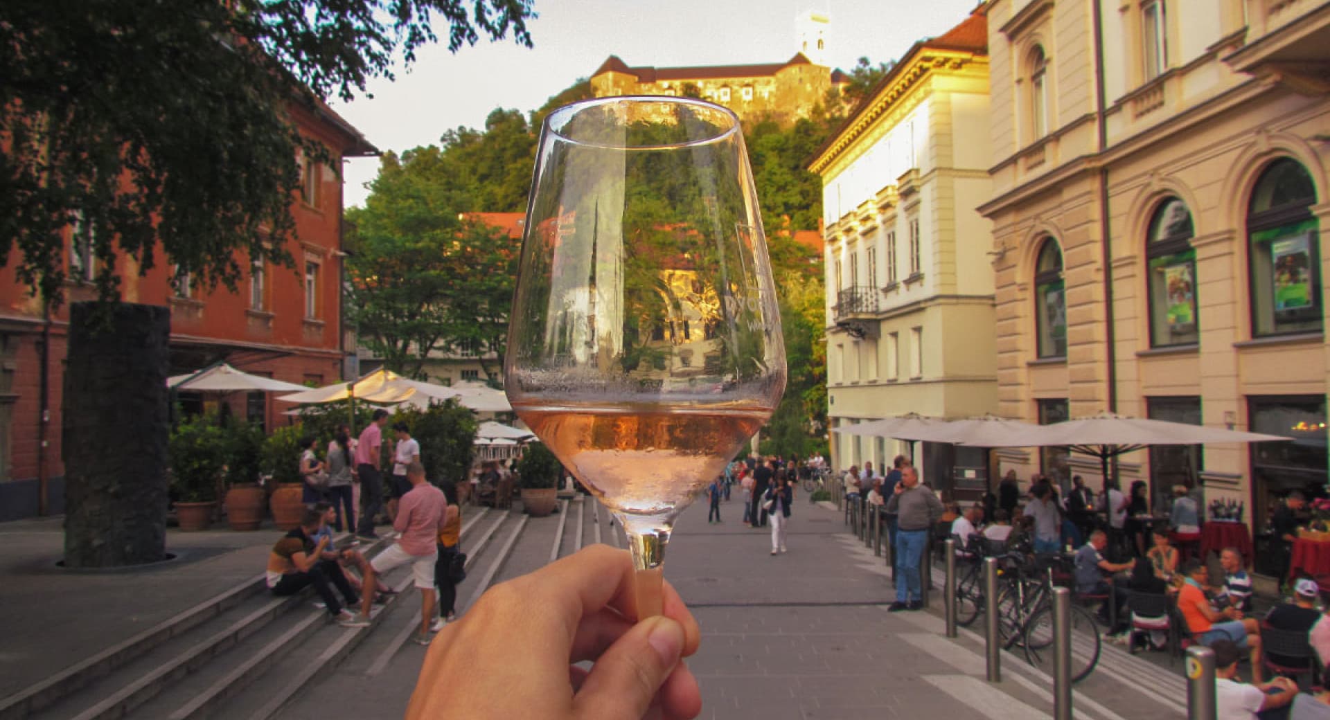 a person holding a glass of wine on the patio outside dvorni wine bar with ljubljana castle in the background slovenia